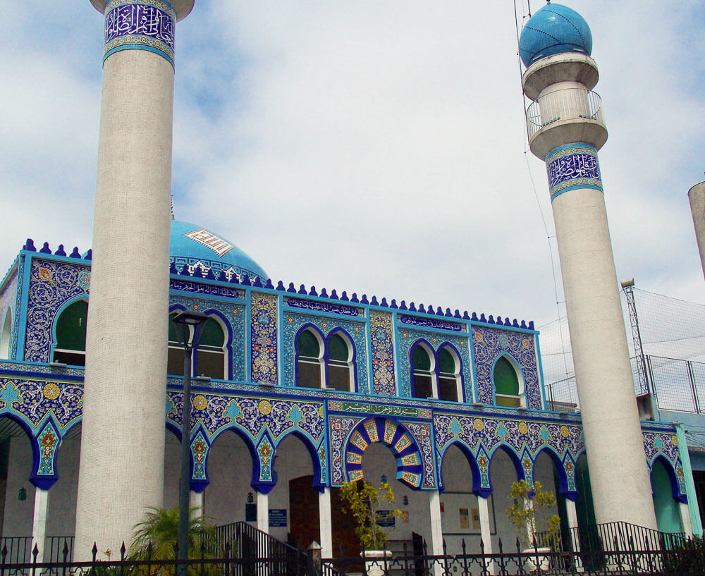 Visão frontal da Mesquita Imam Ali Ibn Abi Tálib, com arabescos azuis, arcos, uma cúpula e duas torres.