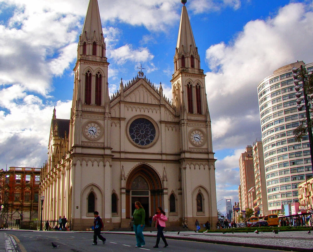 A Catedral de Curitiba vista de frente durante o dia. Há pessoas andando ao redor da Catedral, e alguns prédios aparecem ao fundo.