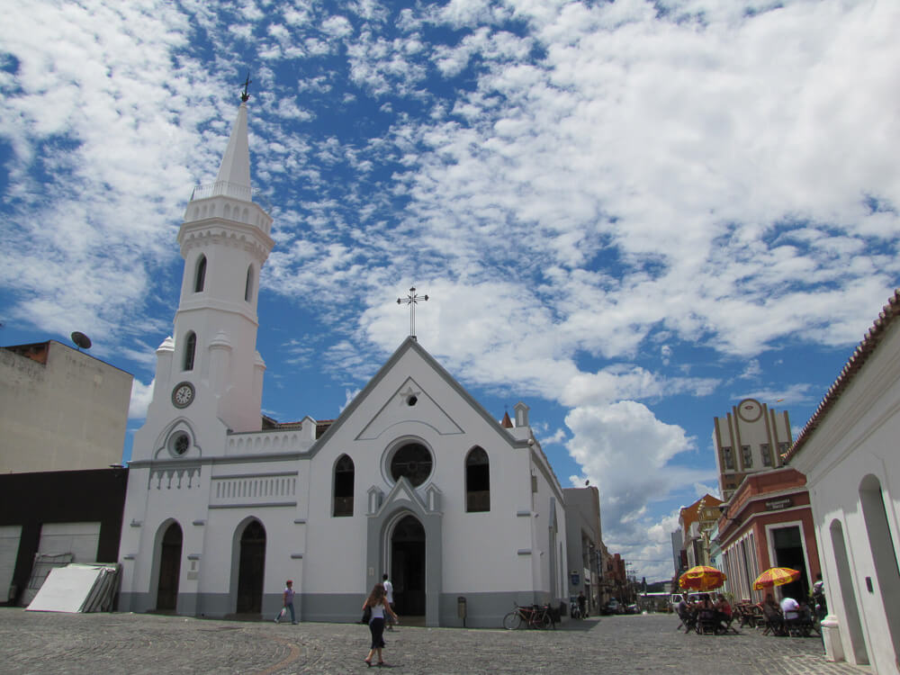 Fachada da Igreja da Ordem, com paredes acinzentadas e, à direita, alguns estabelecimentos comerciais coloridos.