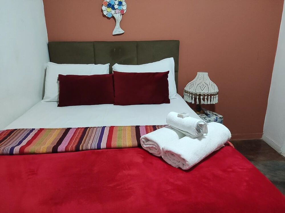 Quarto da Pousada Cantinho da Shirloca, com cama de casal e uma parede em tom terroso.
