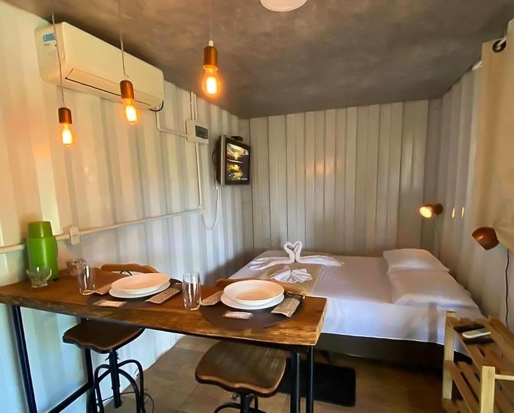 Quarto da Pousada Cipó Container Chalés, com cama de casal, bancada com cadeiras e TV.