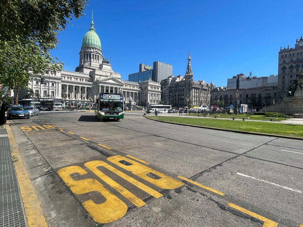 Ônibus portenhos circulando em rua em frente ao Palácio do Congresso, no centro de Buenos Aires, durante o dia.