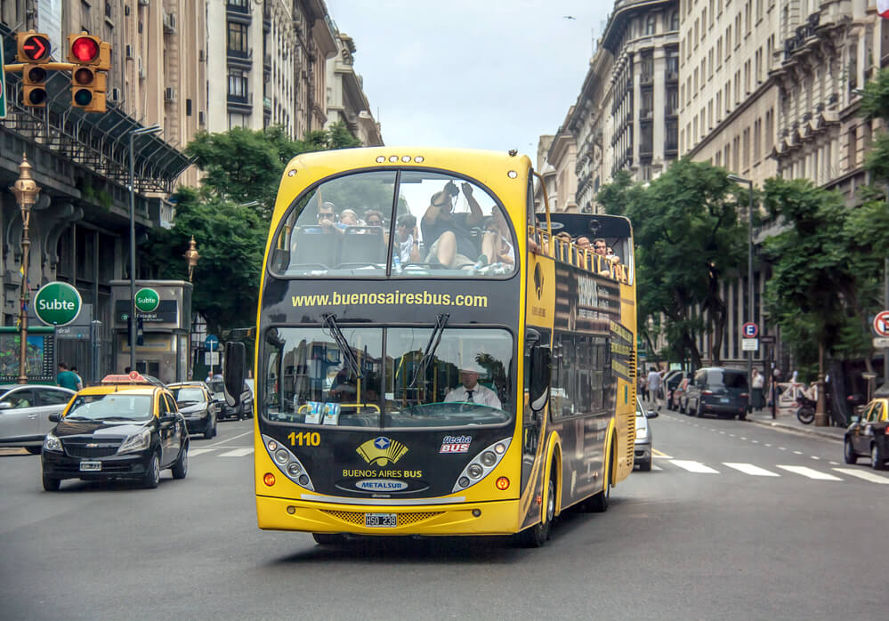 Ônibus turístico de dois andares, da empresa Buenos Aires Bus, circulando durante o dia.