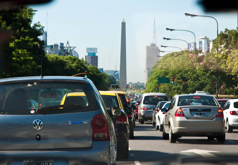 Foto tirada de dentro de carro no trânsito da Avenida 9 de Julio, com visão para o Obelisco de Buenos Aires, durante o dia.