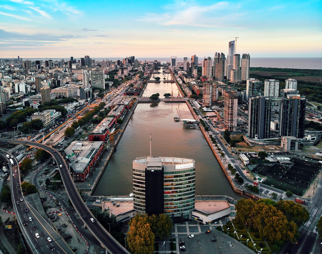 Vista aérea do bairro Puerto Madeiro, em Buenos Aires, durante o dia. A imagem mostra prédios modernos, viadutos, árvores, e pontes e barcos sobre o rio de la Plata.