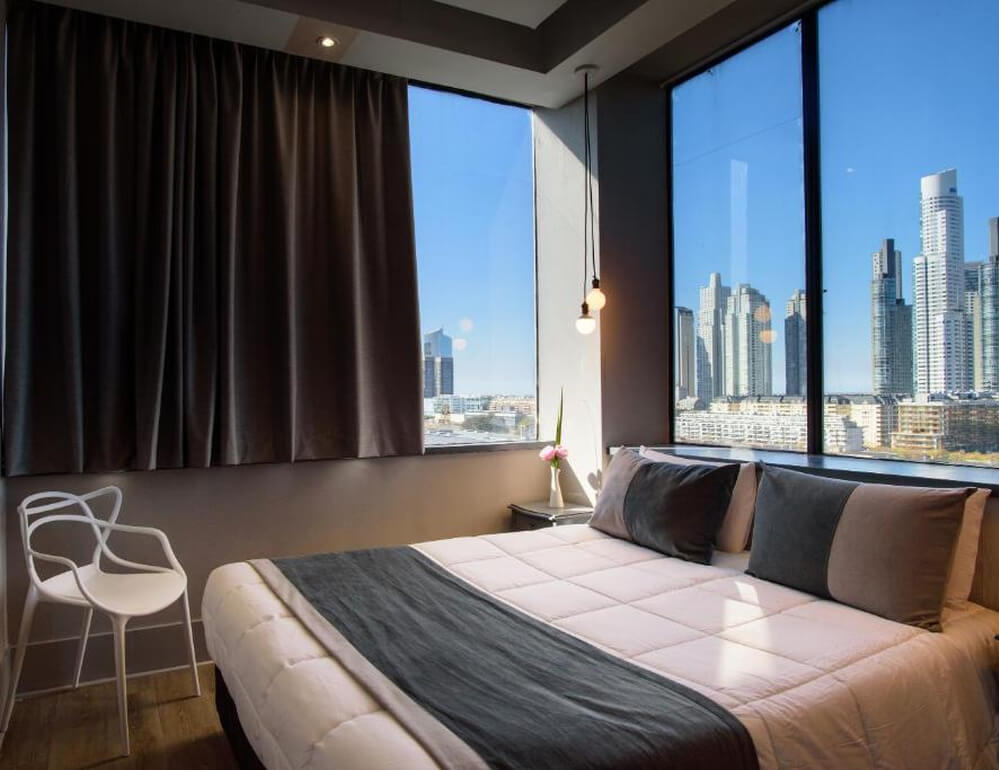 Quarto do hotel Believe Madero Hotel com cama de casal, cadeira e janelas com vista para a cidade.