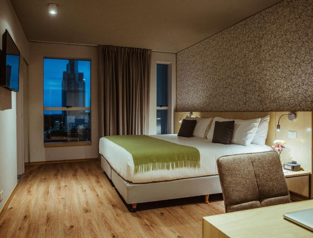 Quarto do hotel Pleno Madero, com cama de casal, tv, e janela com vista para a cidade à noite.