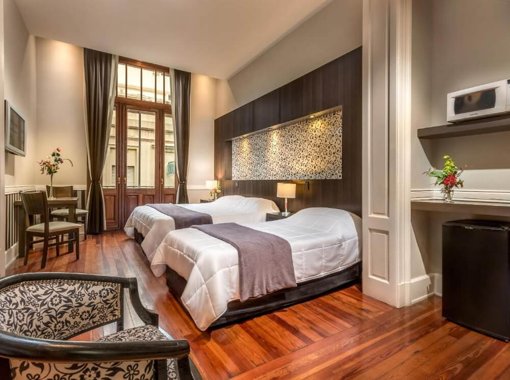 Quarto do hotel Up Tribeca, com uma cama de casal, uma cama de solteiro, microondas, frigobar, poltrona, mesa com cadeiras e varanda.