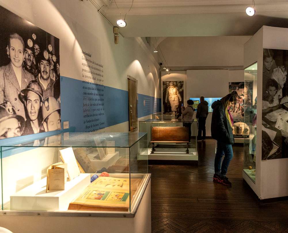 O interior do Museo Evita, com fotografias nas paredes ilustrando ações de Evita Perón, e objetos e documentos expostos através de caixas de vidro transparente.