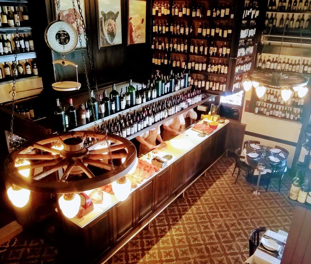 O interior do restaurante Don Julio, com muitas garrafas rotuladas em prateleiras, lustres, uma bancada onde funcionários trabalham, e uma mesa vazia.