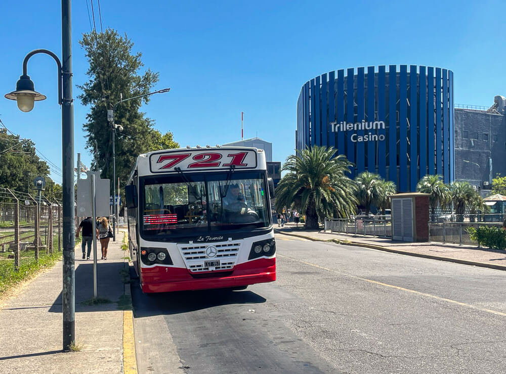 Fachada do Cassino Trilenium com ônibus vermelho ao lado, durante o dia.
