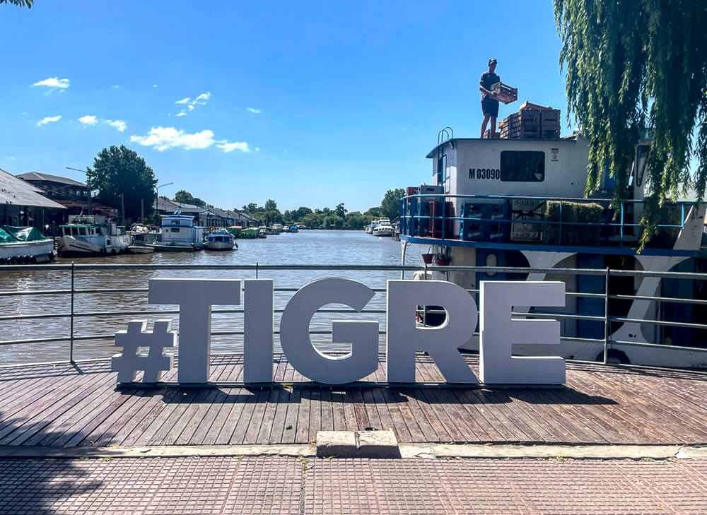 O letreiro da cidade de Tigre, com rio e barcos ao fundo, durante um dia ensolarado.