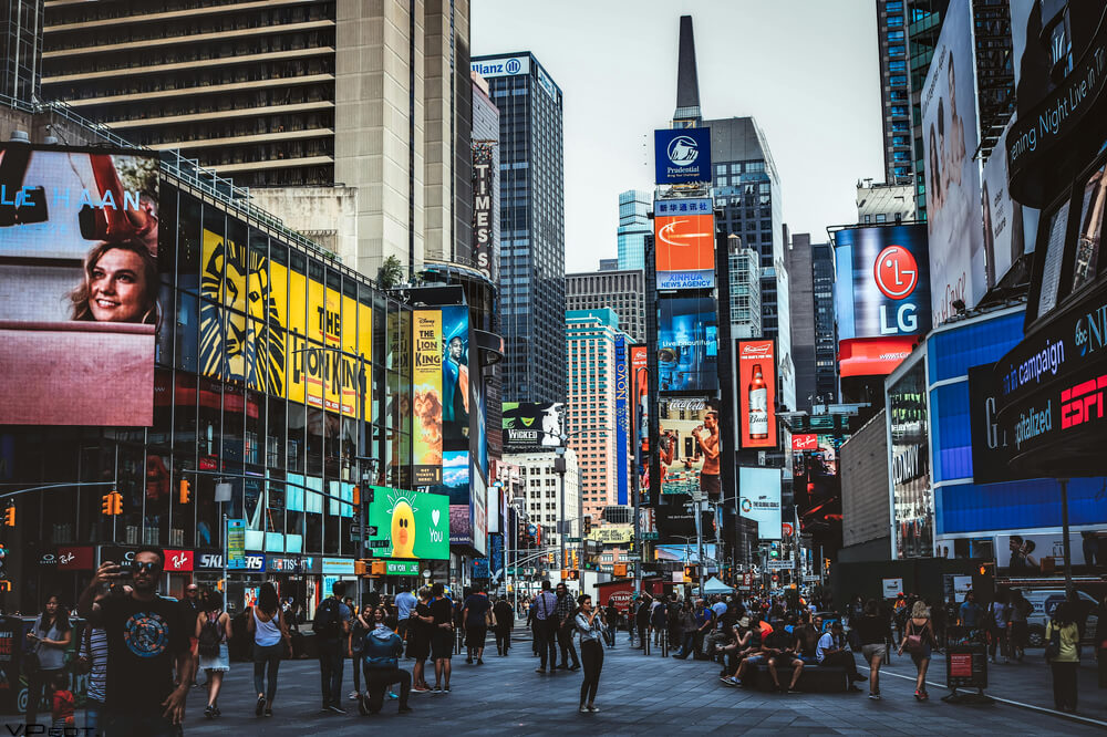 A Times Square durante o dia, há uma multidão de pessoas na rua, anúncios diversos em telões de prédios de diferentes tamanhos.