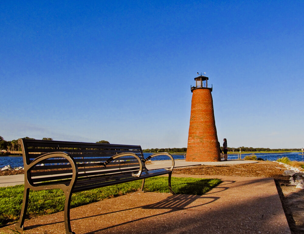 Trecho do parque Lakefront Park em Kissimmee, com banco de madeira, farol e lago, durante o dia.