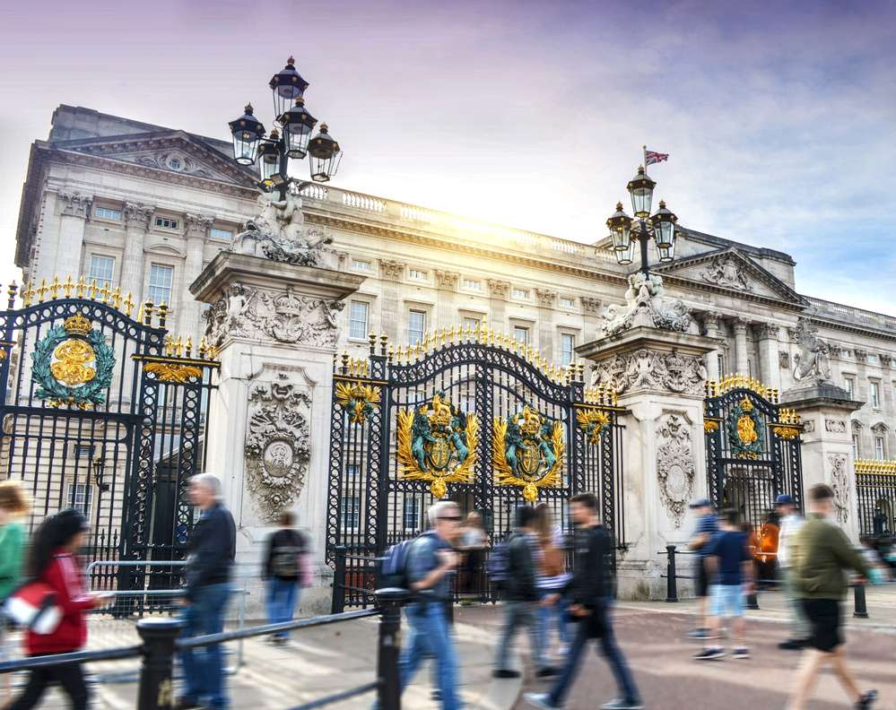 O exterior do Palácio de Buckingham visto durante o dia, através de muros e portões. Pessoas transitam em frente ao local.