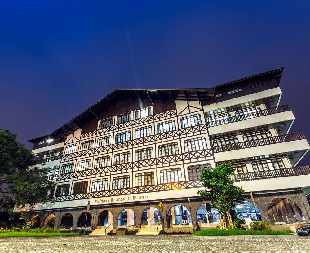 Fachada da Prefeitura Municipal de Blumenau iluminada à noite, destacando a arquitetura enxaimel característica da cidade.