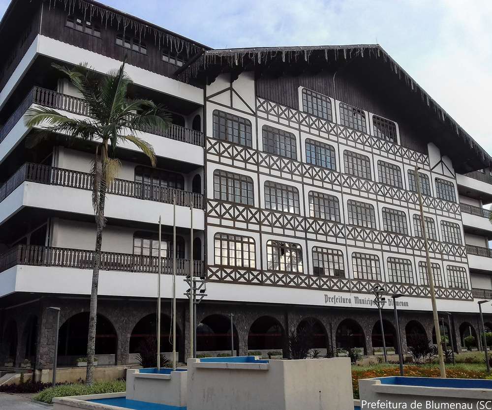 Vista do prédio da Prefeitura de Blumenau, que tem estilo enxaimel. A construção imponente, com detalhes arquitetônicos alemães, é cercada por vegetação.