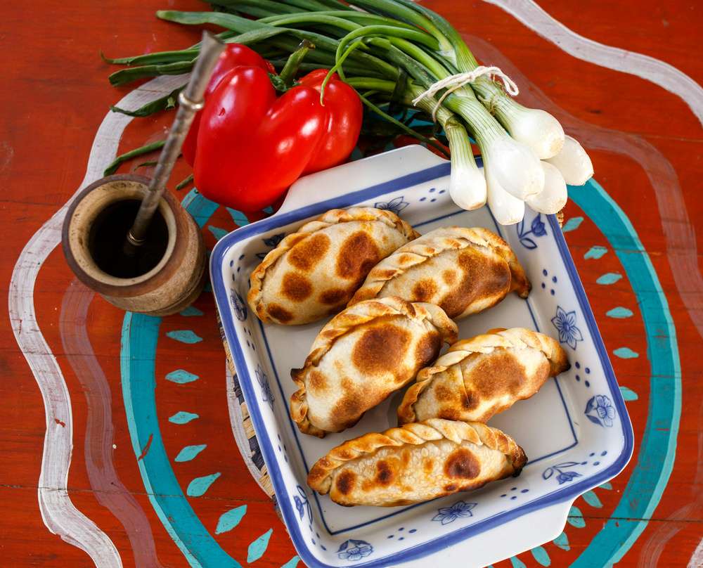Empañadas, mate, pimentão e cebolinhas sobre uma mesa colorida em vermelho e azul.