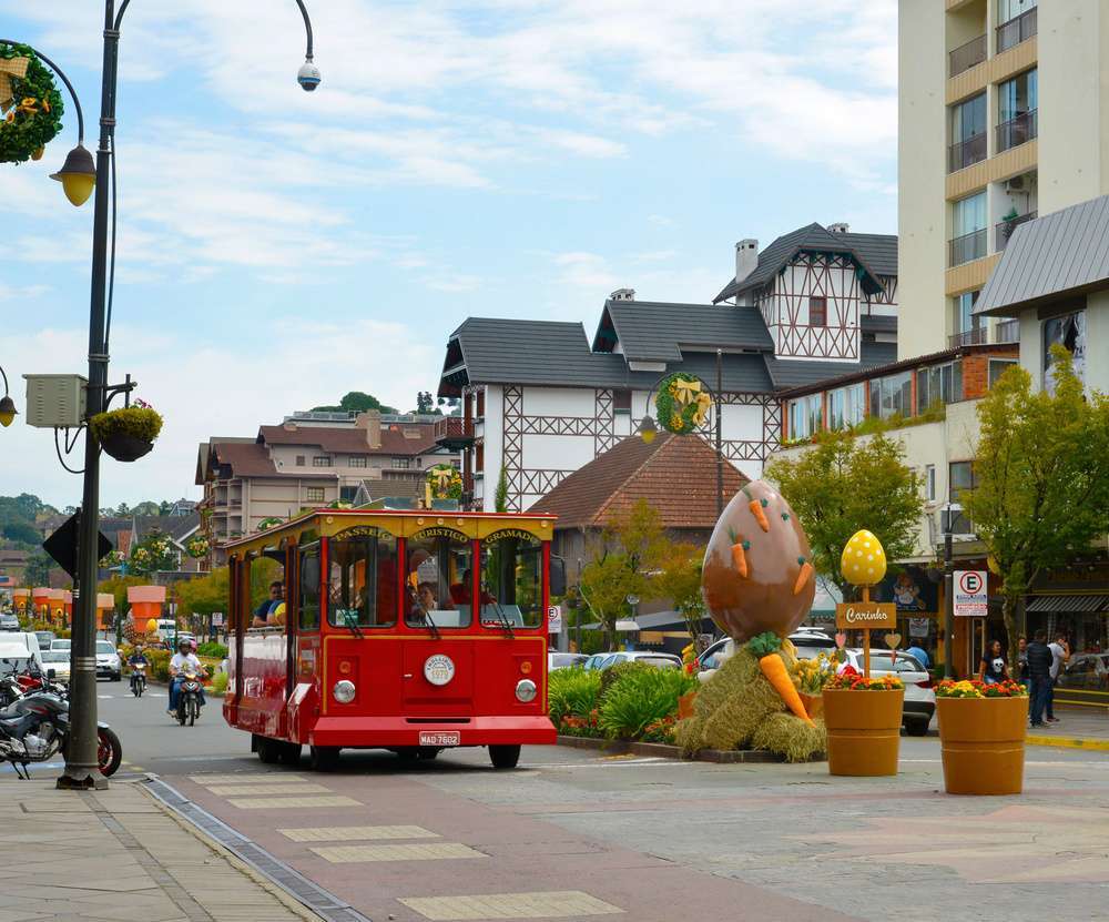 A Jardineira de Gramado, um bonde turístico vermelho circulando por rua de Gramado decorada para a Páscoa, com edifícios em estilo europeu ao fundo.