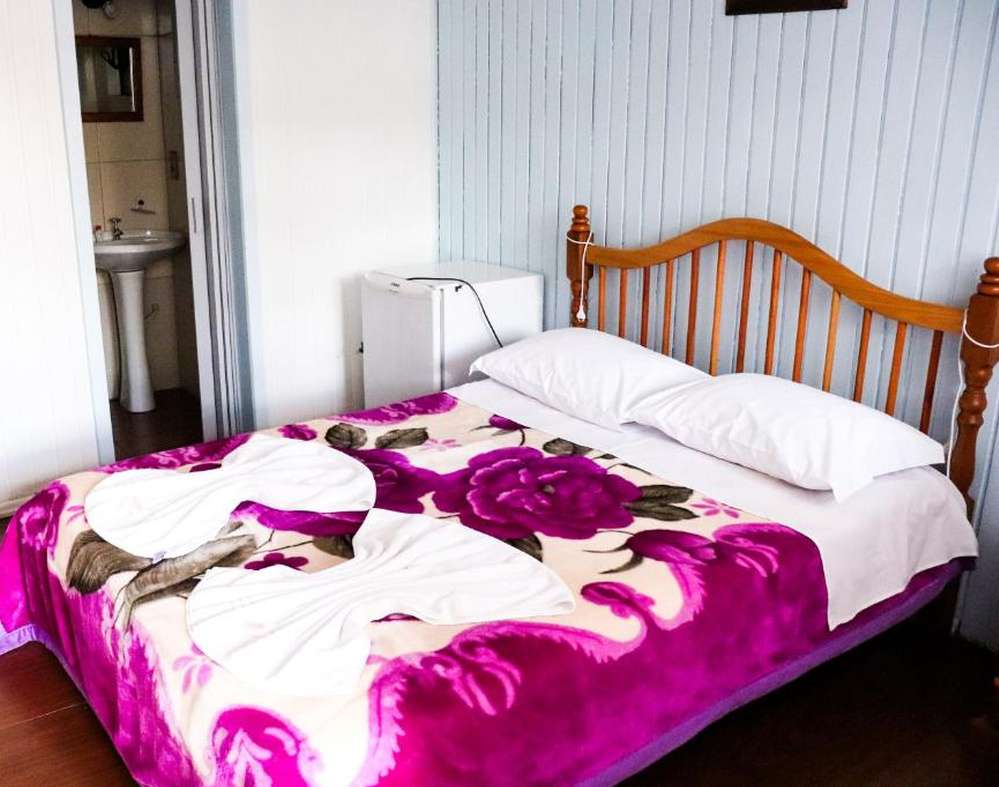 Quarto da Pousada Tia Leonor com cama de casal, cobertor floral roxo, frigobar, banheiro com pia e espelho ao fundo.