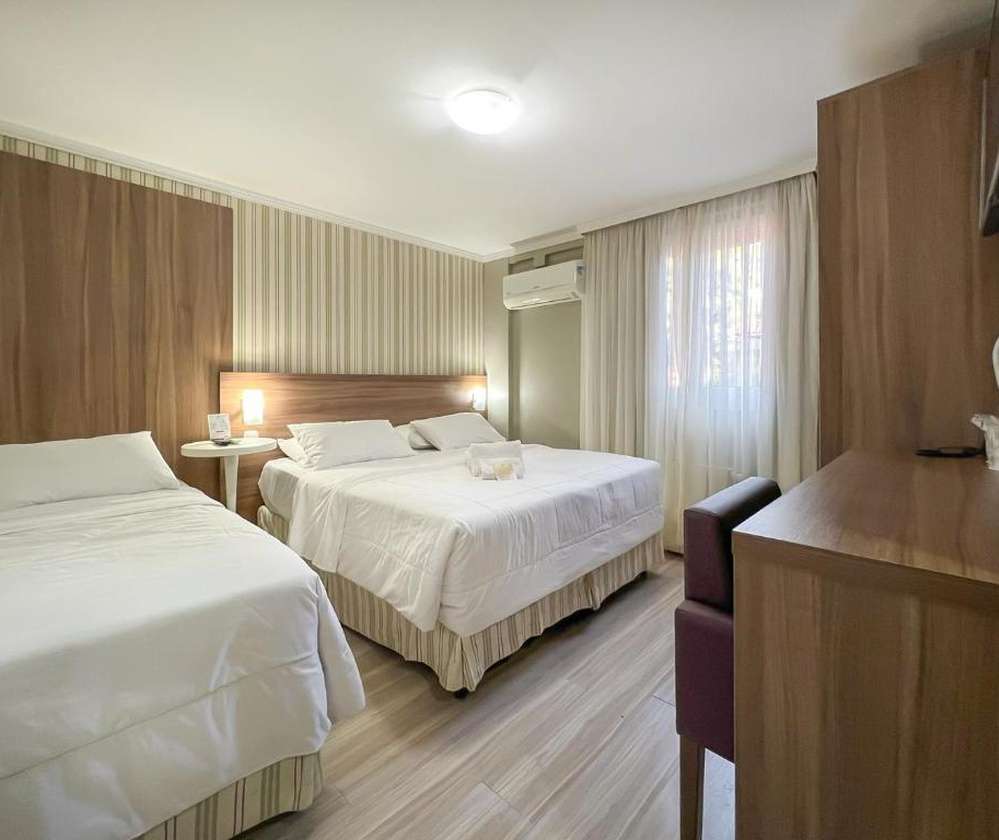 Quarto da Pousada Viena em Gramado, com duas camas de casal, decoração em tons claros e madeira, mesa lateral, ar-condicionado, cortinas brancas e uma pequena mesa de trabalho.