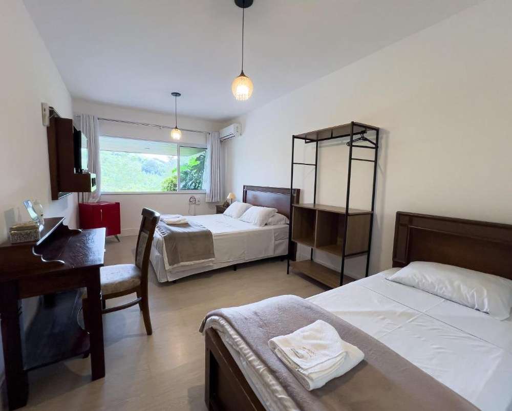 Quarto da Casa Branca Pousada, com uma cama de casal e uma de solteiro, decoração simples e móveis de madeira, com uma grande janela oferecendo vista para a vegetação.