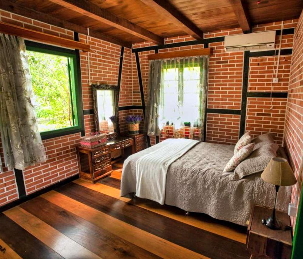 Quarto da Pousada Imigrante, com cama de casal, paredes de tijolos vermelhos, e janela com vista para vegetação.