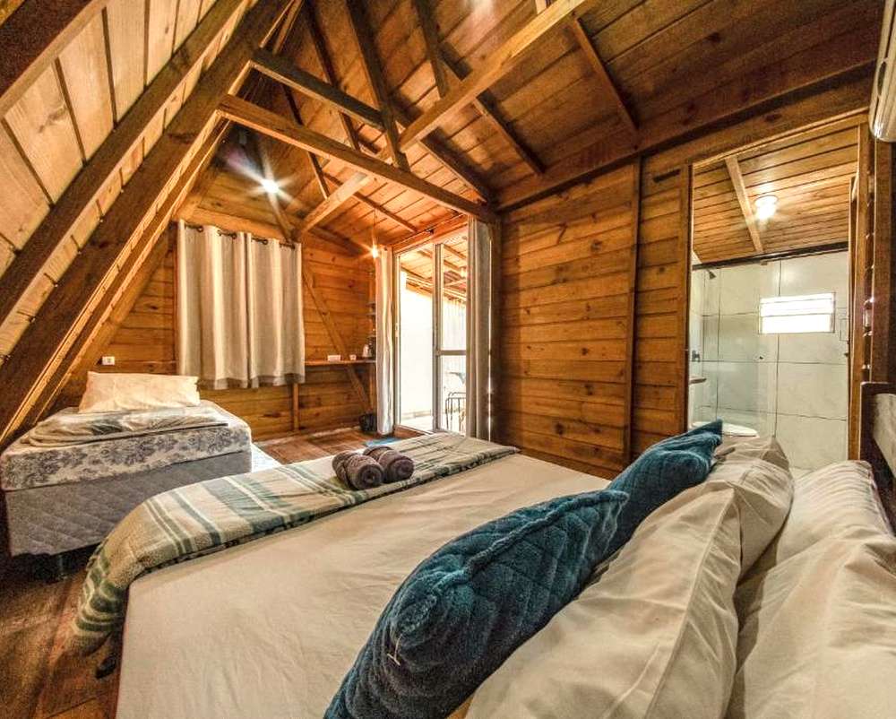 Quarto aconchegante no Recanto do Vale em Pomerode, com cama de casal, roupa de cama branca, abajur, criado-mudo, cortinas bege e janela ampla.