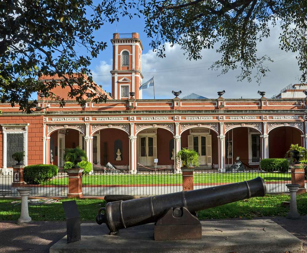O Museo Histórico Nacional em Buenos Aires, com fachada vermelha, varanda decorada e canhões expostos no jardim da frente.