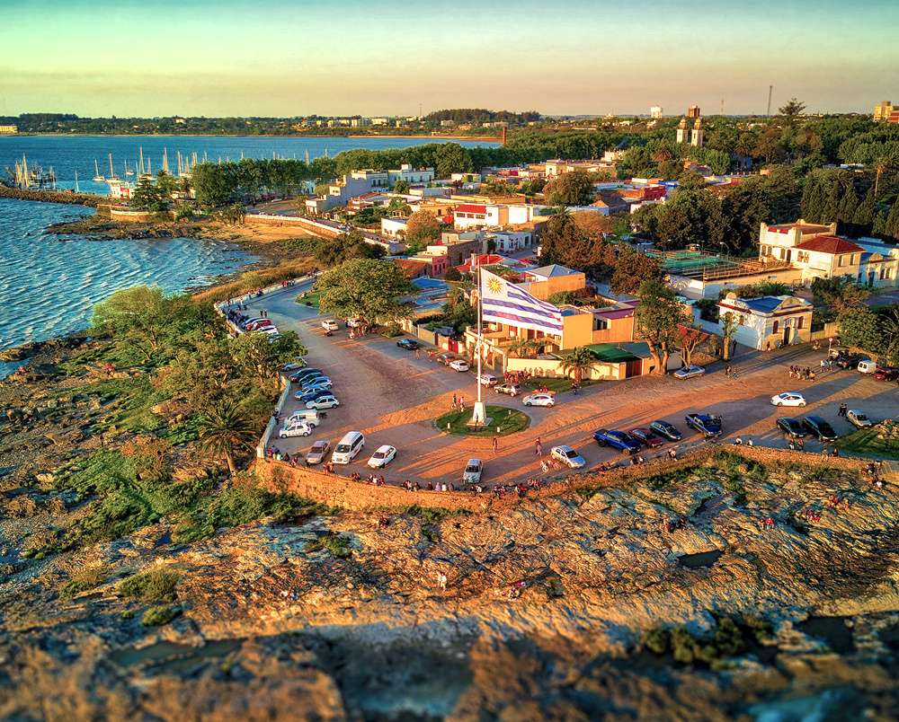 Vista aérea do Bastión de San Pedro em Colonia del Sacramento, ao pôr do sol, com bandeira do Uruguai hasteada, carros estacionados e marina ao fundo.