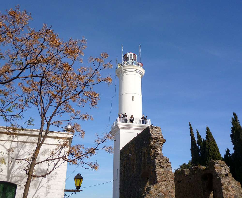 Farol histórico em Colonia del Sacramento em um dia ensolarado, com turistas apreciando a vista do alto.