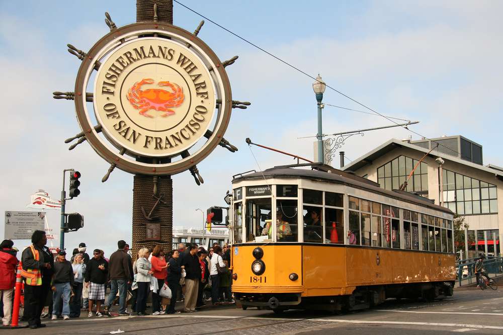 O famoso bondinho amarelo de San Francisco passa pelo movimentado Fisherman's Wharf, com uma multidão de turistas ao fundo.