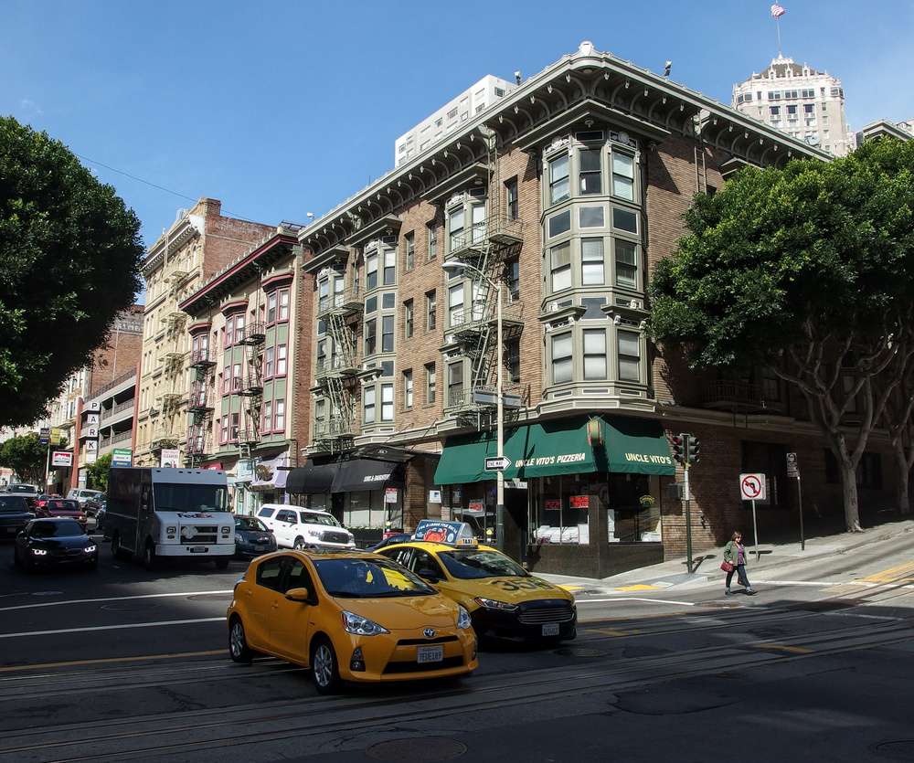 Vista de uma esquina movimentada no bairro de Nob Hill, em San Francisco, com prédios históricos, um restaurante e veículos passando pela rua.