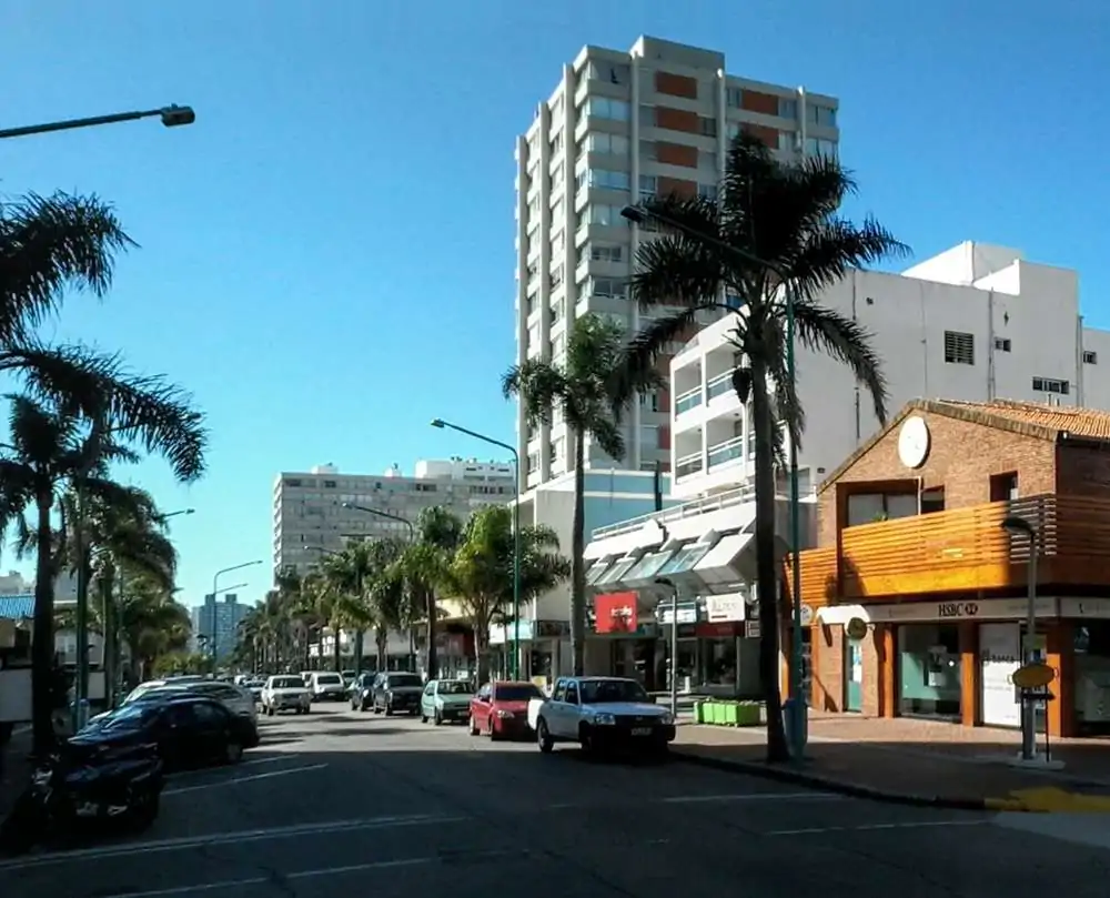 A movimentada Avenida Gorlero em Punta del Este, com prédios altos, palmeiras e lojas. O dia está ensolarado, criando uma atmosfera agradável para passear pela principal via da região da Península.