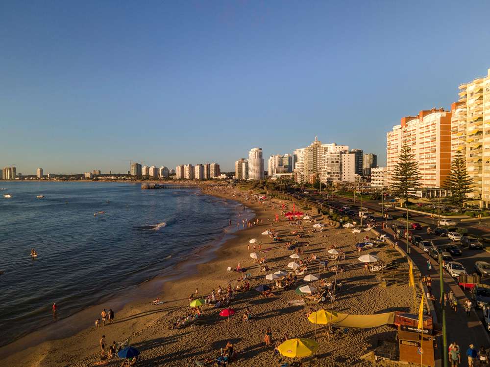 Vista aérea da Playa Mansa, em Punta del Este, com edifícios modernos ao fundo e pessoas aproveitando o pôr do sol na areia.