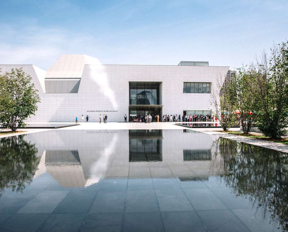 Fachada moderna do Museu Aga Khan, em Toronto, refletida em um espelho d'água, com visitantes entrando no edifício minimalista de paredes brancas.