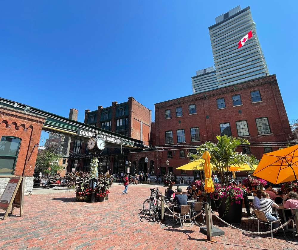 Turistas relaxam em um charmoso café ao ar livre no Distillery District, Toronto, rodeados por edifícios históricos de tijolos vermelhos e o famoso relógio Gooderham & Worts, com um céu azul como pano de fundo.