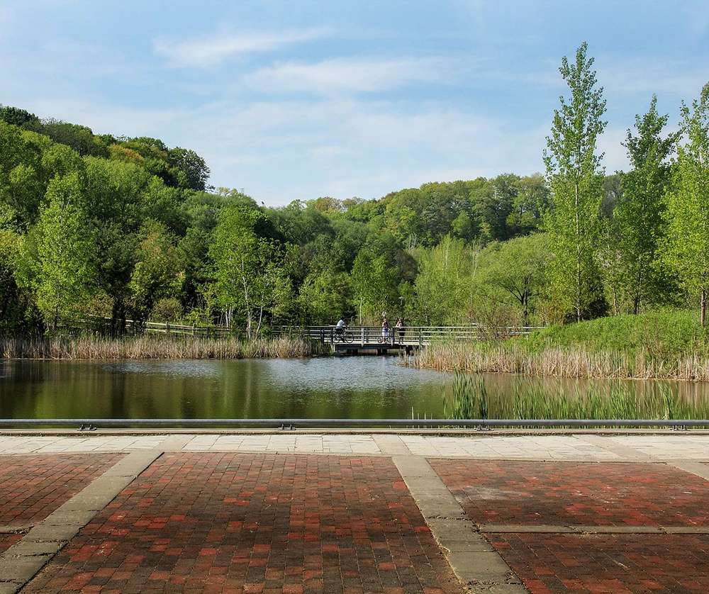 Vista de um lago cercado por árvores no Evergreen Brick Works, com pessoas passeando em uma ponte ao fundo.