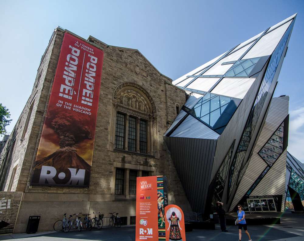 A fachada do Museu Real de Ontário com uma mistura de arquitetura antiga e moderna, destacando banners de exposições sobre Pompeia e a cultura mexicana.