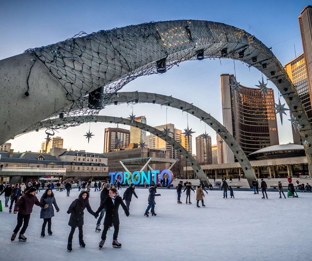 Pessoas patinando no gelo em frente ao famoso letreiro colorido de Toronto, na Nathan Phillips Square, cercados por arcos iluminados e arranha-céus ao fundo.