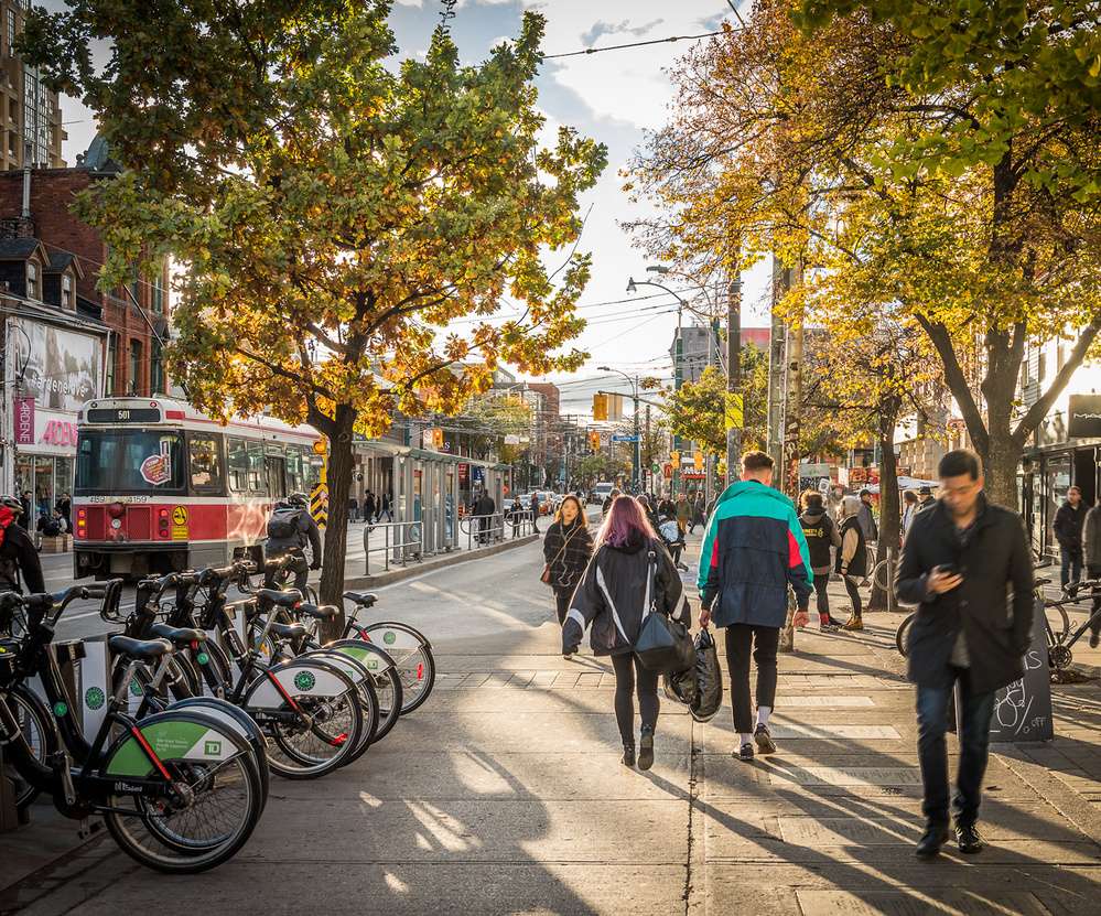 Pessoas caminhando na movimentada Queen Street em Toronto, ao lado de um ponto de bicicletas compartilhadas, com árvores ao fundo e um bonde vermelho à vista.