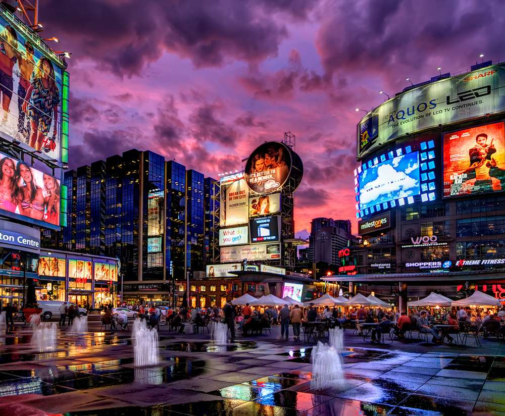 Uma noite vibrante na Yonge-Dundas Square, em Toronto, com luzes de letreiros e telões iluminando a praça cheia de pessoas.