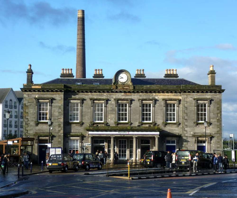 Fachada histórica da estação ferroviária de Haymarket em Edimburgo, com táxis estacionados e pessoas caminhando em frente ao edifício.