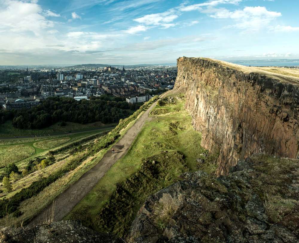 Vista do Holyrood Park em Edimburgo, com penhascos e trilhas, e a cidade ao fundo em um dia ensolarado.
