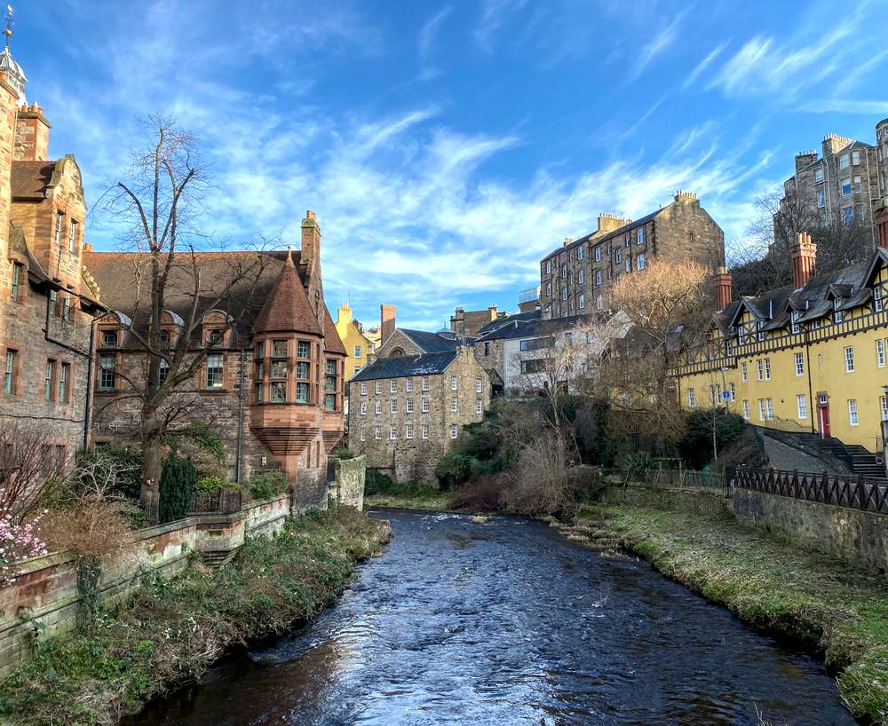 O vilarejo Dean Village em Edimburgo, com casas coloridas e tradicionais à beira de um rio tranquilo, em um dia claro e ensolarado.