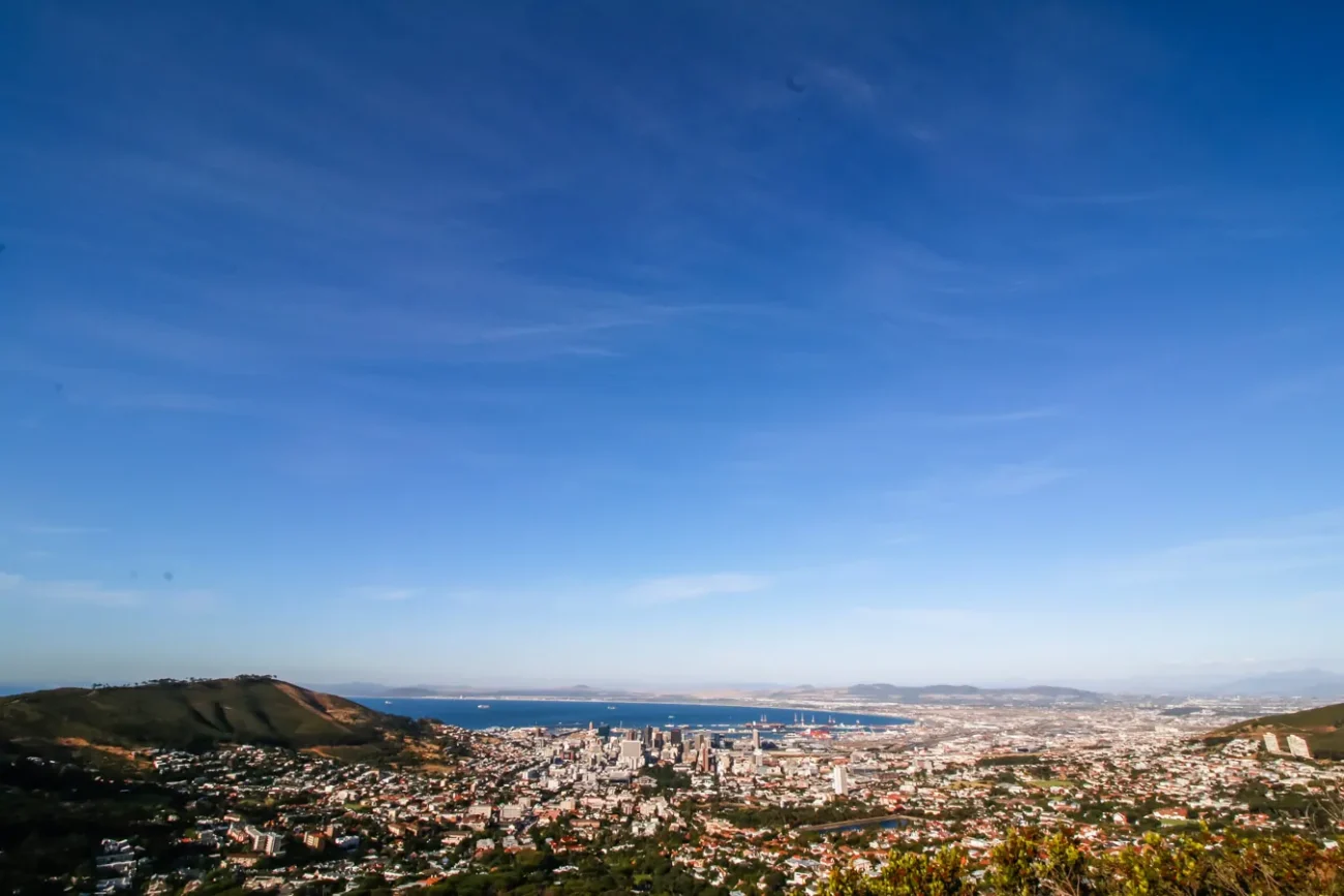 Vista aérea da cidade de Cape Town com céu claro e o mar ao fundo. A cidade se estende ao longo da baía, cercada por colinas verdes.