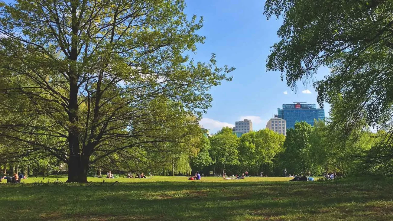 Área bastante arborizada em Tiergarten, com várias pessoas descansando sob a sombra das árvores e alguns edifícios ao fundo.