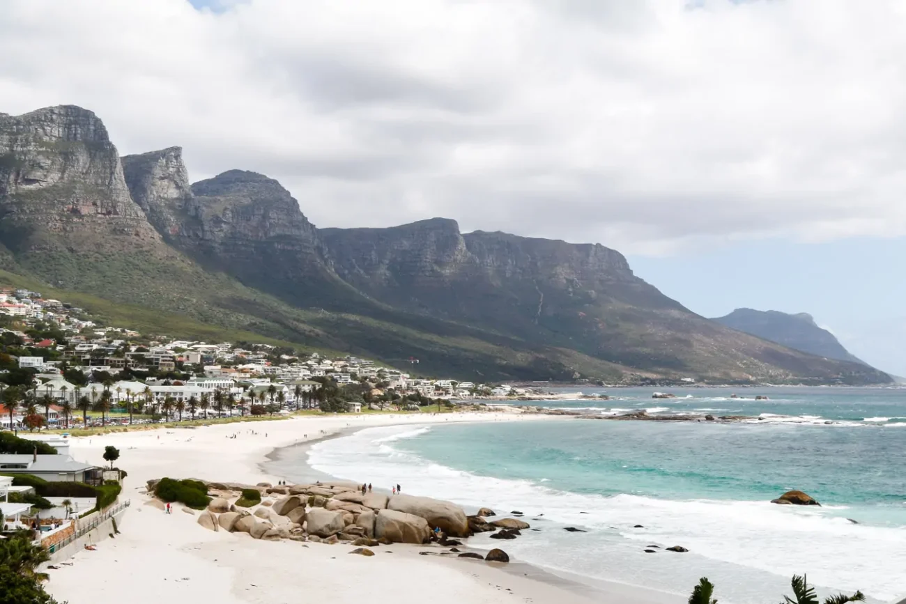 Vista panorâmica de Camps Bay, Cape Town, com uma praia de areia branca e o mar azul ao fundo. As montanhas da região cercam a área, e há várias casas estão distribuídas pela encosta.