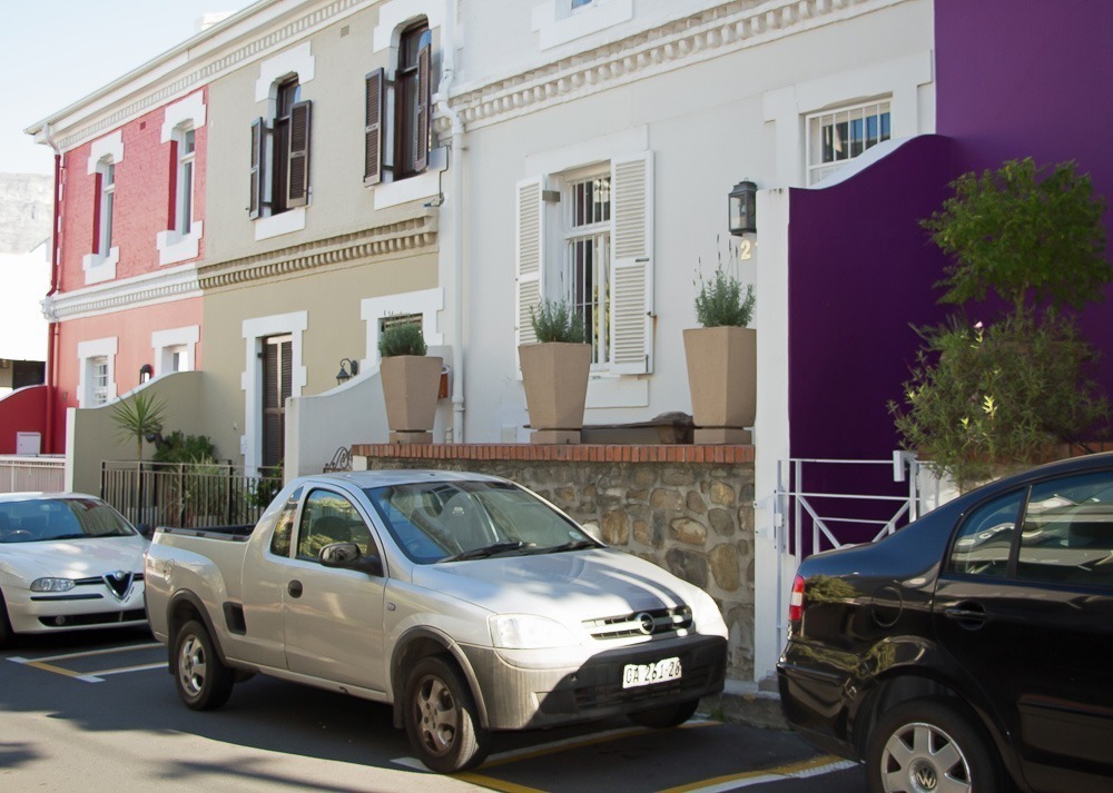 Fachadas de casas no bairro De Waterkant, em Cape Town, com tons de vermelho, bege e branco, além de um toque vibrante de roxo em uma parede lateral.