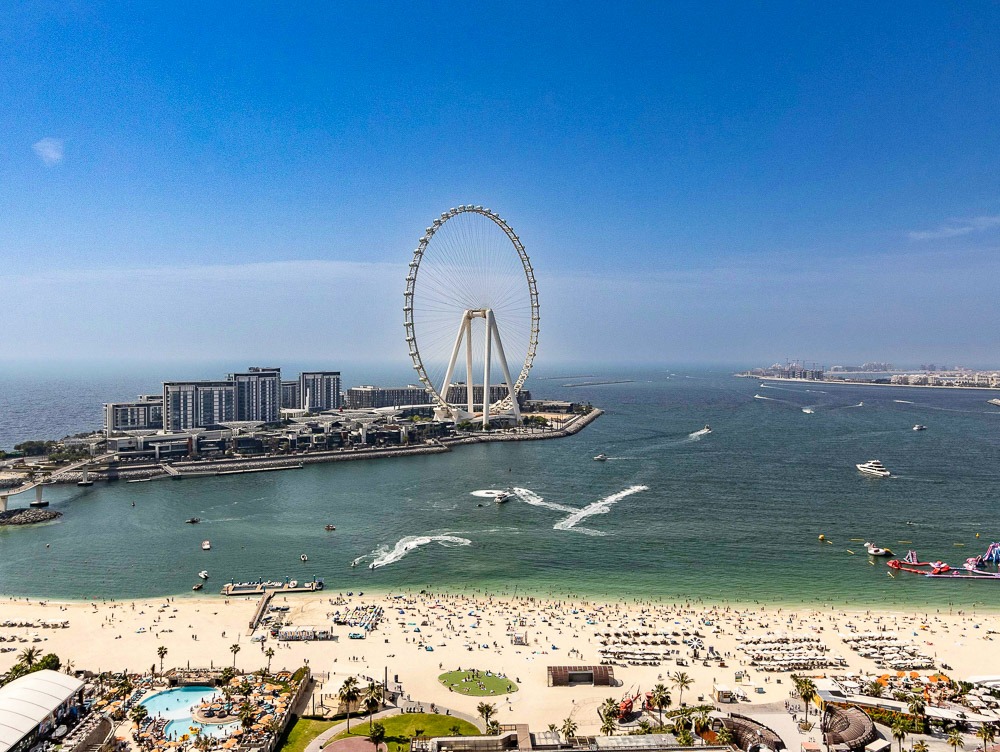 A roda-gigante Ain Dubai vista de longe e a Marina de Dubai, com uma areia extensa e repleta de banhistas.